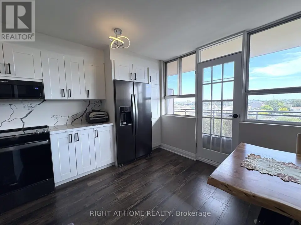 Dining area with wood table beside windows and glazed balcony door; refrigerator and range at left; neighbourhood view outside.