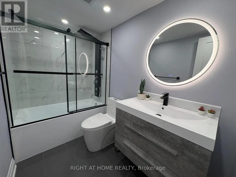 Bathroom with glass-enclosed tub, black shower panel, white toilet, grey vanity and round backlit mirror.