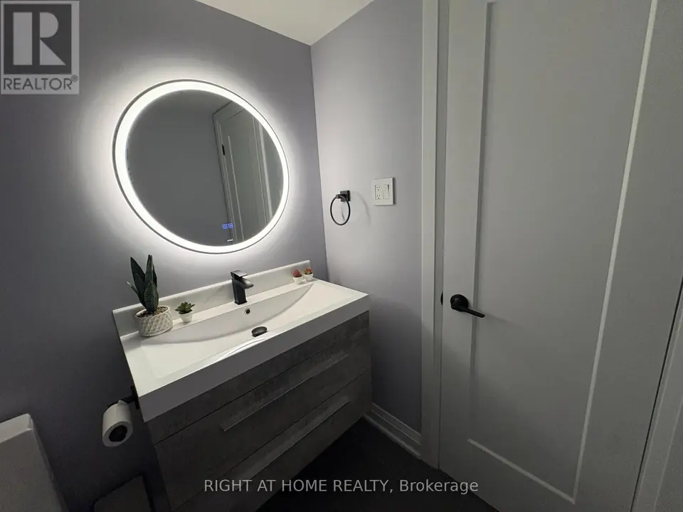Grey vanity with white sink and black faucet under a round backlit mirror, beside a white door.
