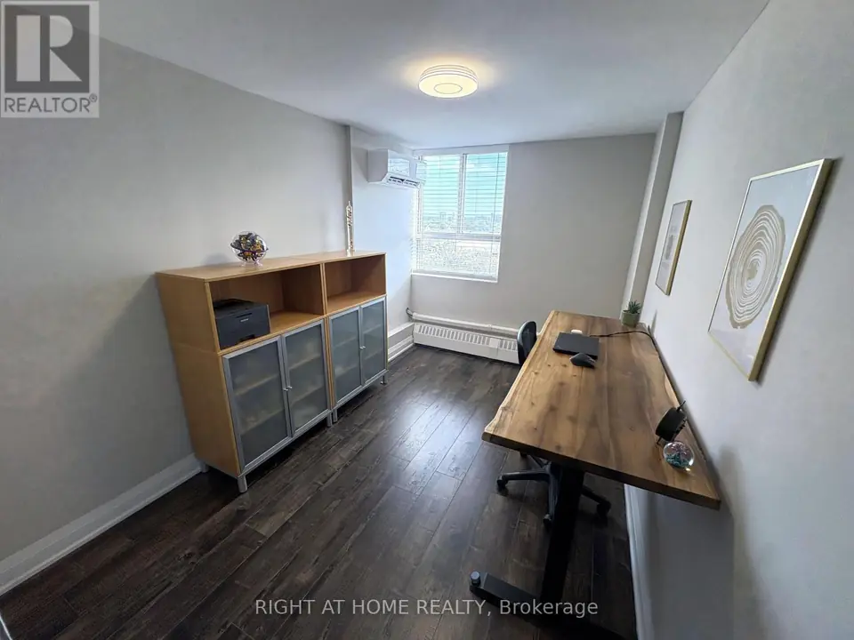 Small bedroom set up as an office with a wooden desk, chair, two storage cabinets and a window with blinds.