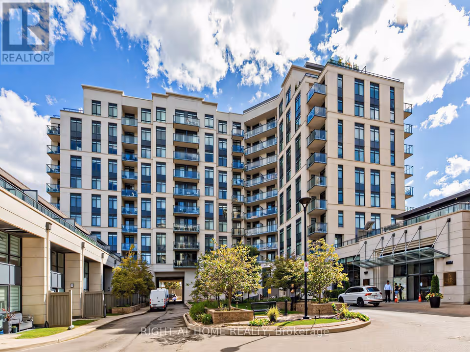 A cream-coloured multi-storey condominium building with rows of glass-railed balconies rises behind a courtyard of trees and planting beds, with a curved driveway in front. At right, a glass canopy shelters the entrance, with a white SUV and a few small figures beside it, under a blue sky with bright clouds.
