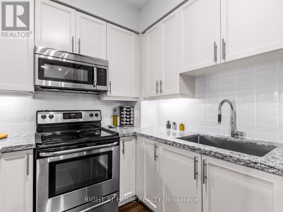 A close view of the kitchen corner: a stainless steel range with a smooth cooktop sits under an over-the-range microwave, with a spice rack and a bottle on the speckled granite counter beside it. At right, a stainless steel sink with a pull-down faucet sits below lit white cabinets and a white tile backsplash.
