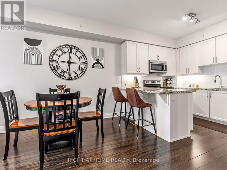 A round wooden dining table with black chairs stands in the left foreground, with a large black Roman-numeral wall clock and two graphic art prints on the wall behind it. To the right, a granite-topped breakfast bar with two brown upholstered stools fronts a white kitchen with a stainless steel microwave and range, under-cabinet lighting and a tiled backsplash, all over dark laminate flooring.