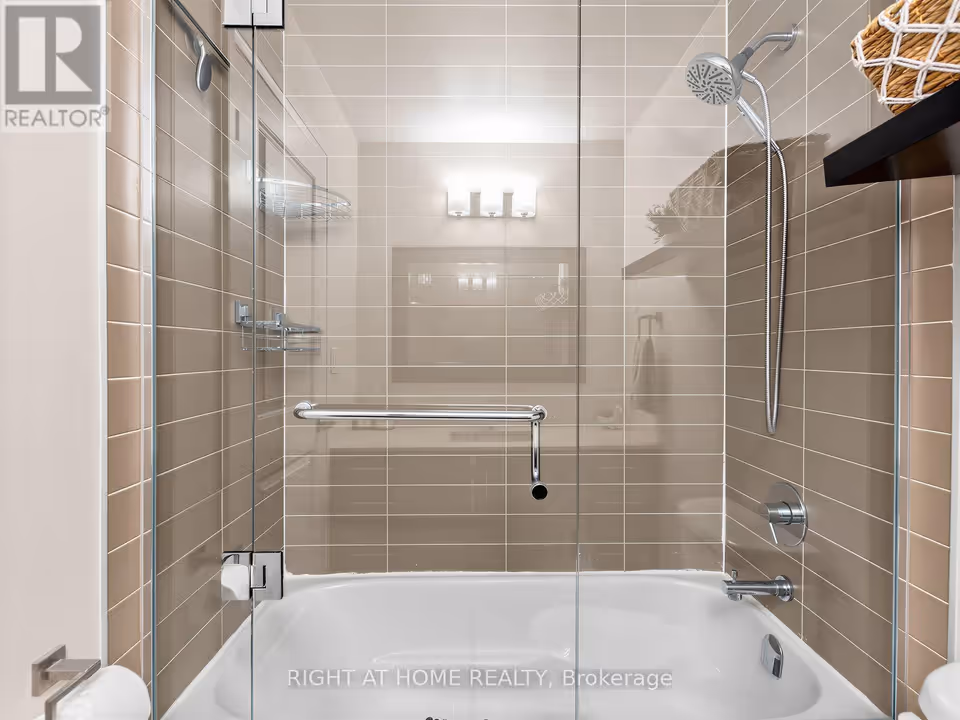 Straight-on view of a white bathtub enclosed by a frameless glass door with a chrome bar handle. The recess is tiled in horizontal taupe rectangles, with a handheld shower head and hose on the right wall, a chrome tub spout and lever control, and wire shower caddies at left.