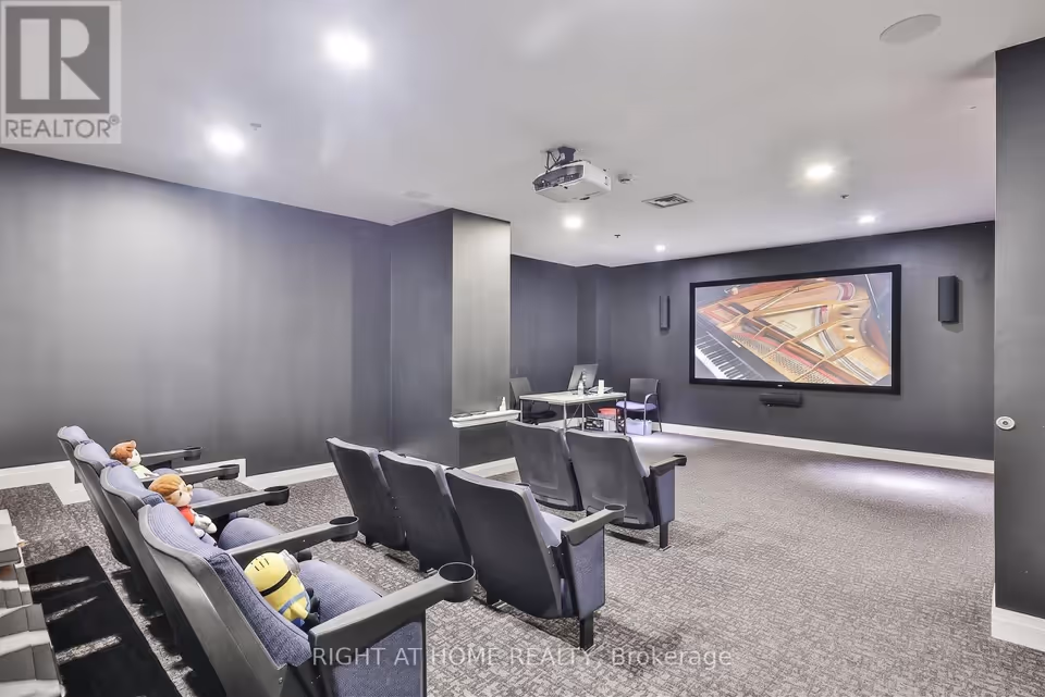 A screening room with dark grey walls and grey patterned carpet. Dark cinema-style seats, some holding plush toys, face a large framed screen showing a piano interior, with a ceiling projector above, speakers on either side of the screen and a small desk with a monitor at the back.